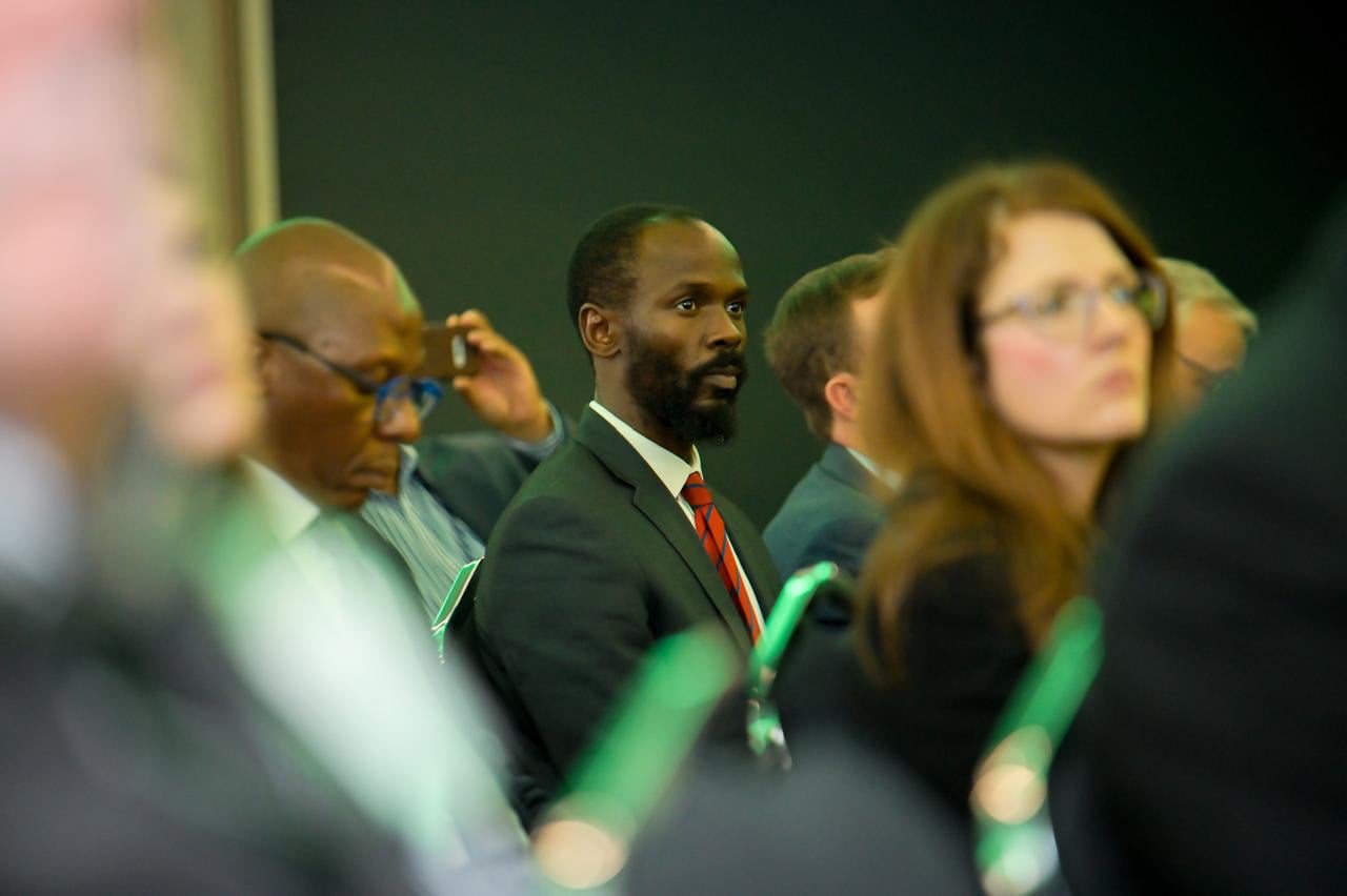 A man in a dark suit sits attentively among an audience at an industry session, other attendees softly out of focus around him.