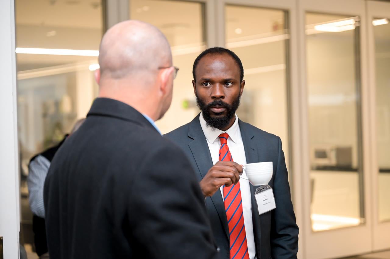 Two men in conversation at a professional event; one, in a dark suit and red striped tie, holds a cup and listens closely.