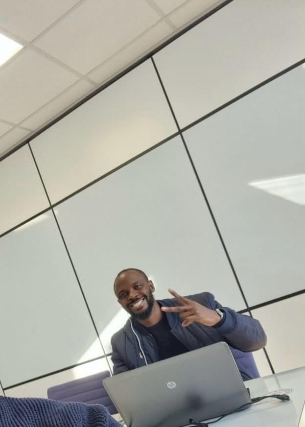 A smiling man at a laptop in a bright meeting room gestures towards the camera.